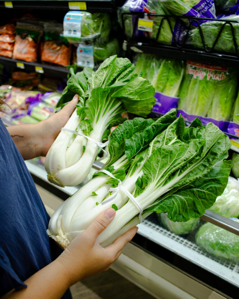 Person holding two bundles of fresh bok choy in the produce section of a grocery store.