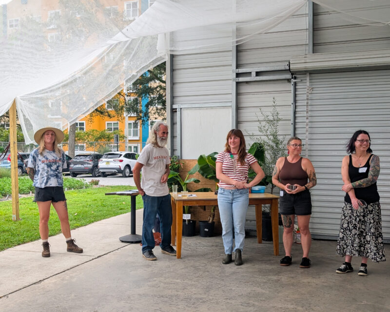 Five people stand in front of a warehouse under a canopy, smiling with plants and a table behind them.