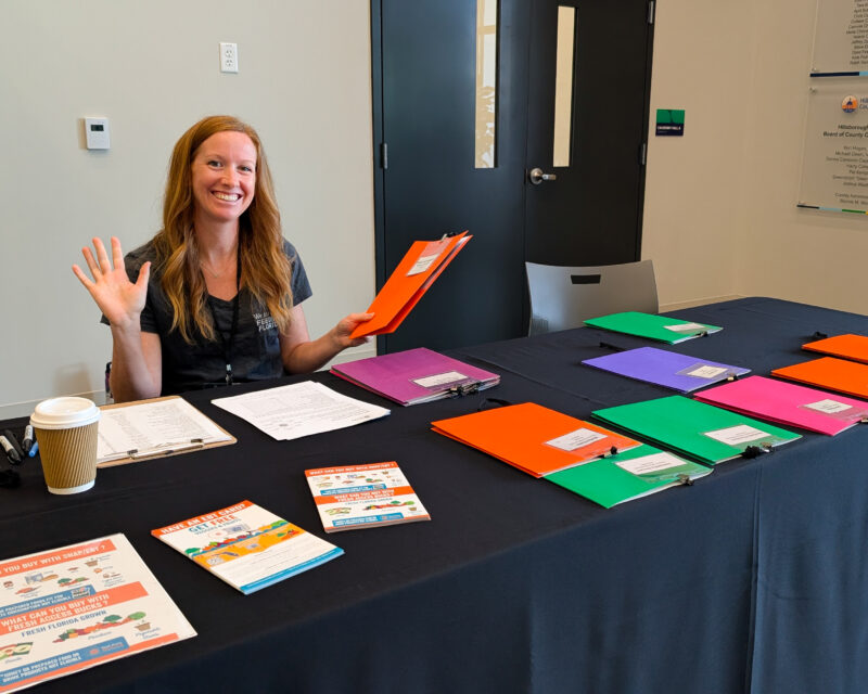 A woman sits at a welcome table with colorful folders, greeting attendees at a conference.