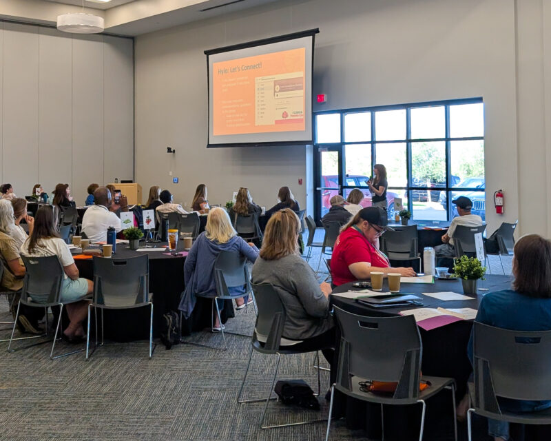 A woman presents at a conference, gesturing to a screen as attendees listen from round tables.