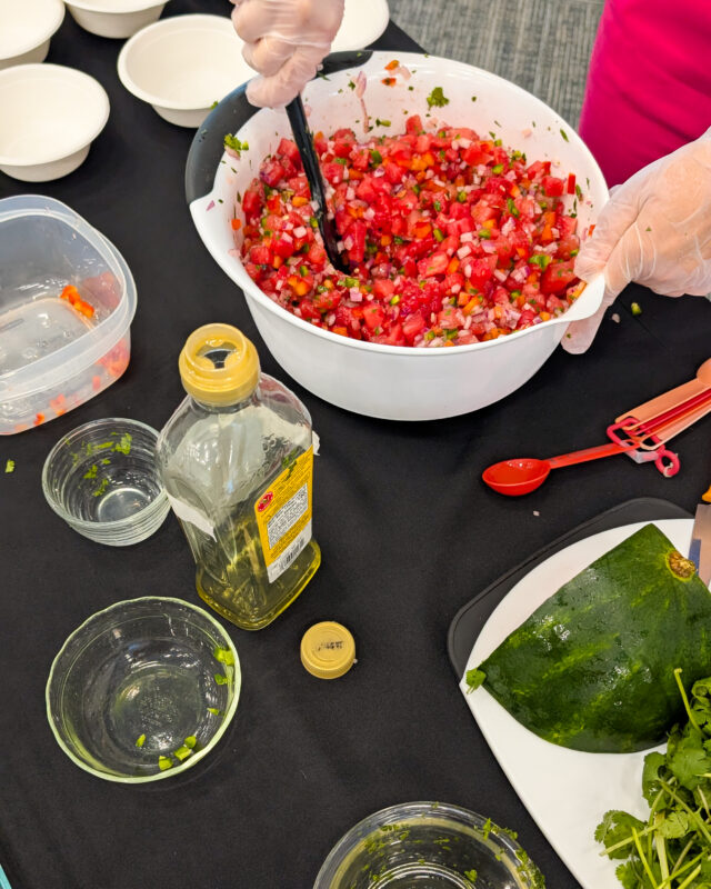 A person prepares watermelon salsa at a cooking demonstration, standing behind a table filled with prepped ingredients. The table holds bowls of cubed watermelon, chopped peppers, diced onion, lemon juice, and a bottle of oil. All ingredients are already cut and arranged for easy mixing. The person is mid-motion, possibly stirring or explaining the process, with a focus on showcasing fresh, colorful, seasonal ingredients.