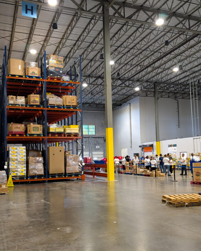 A large group of volunteers wearing white shirts are organizing and packing food in a warehouse. Tall shelves stacked with boxes and food supplies rise in the background, filling the space with abundant donations.