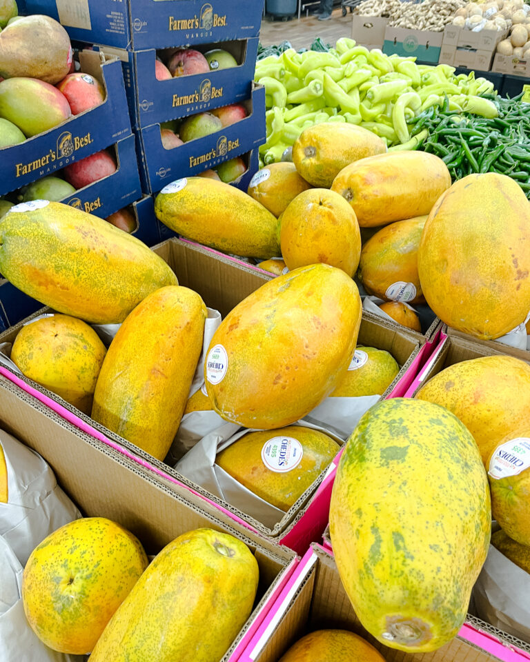Woodman's Papapya (1) Fresh papaya in cardboard boxes next to green peppers and boxes of Farmer’s Best mangos in a grocery produce section.